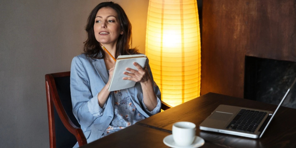 A woman sits at a table, working on a laptop with a cup of coffee beside her.